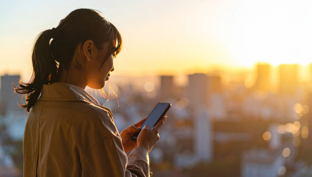 スマートフォンを見る女性、夕日の街並み